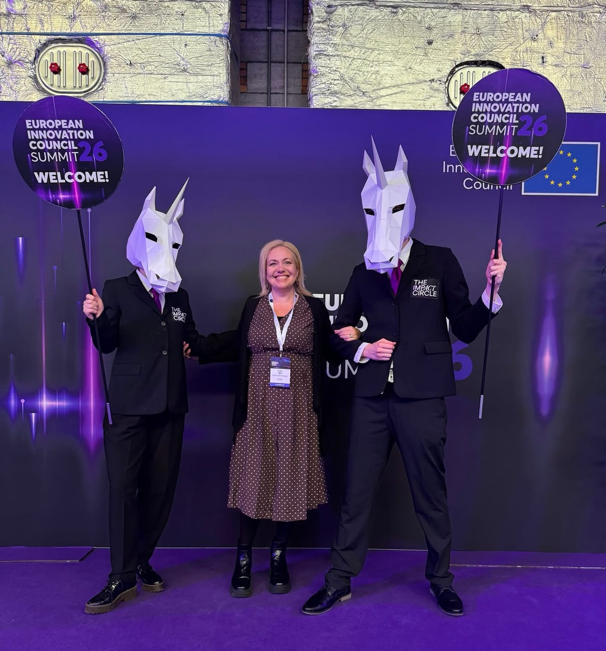 Woman posing between two people in unicorn masks holding European Innovation Council Summit 26 Welcome signs in Brussels